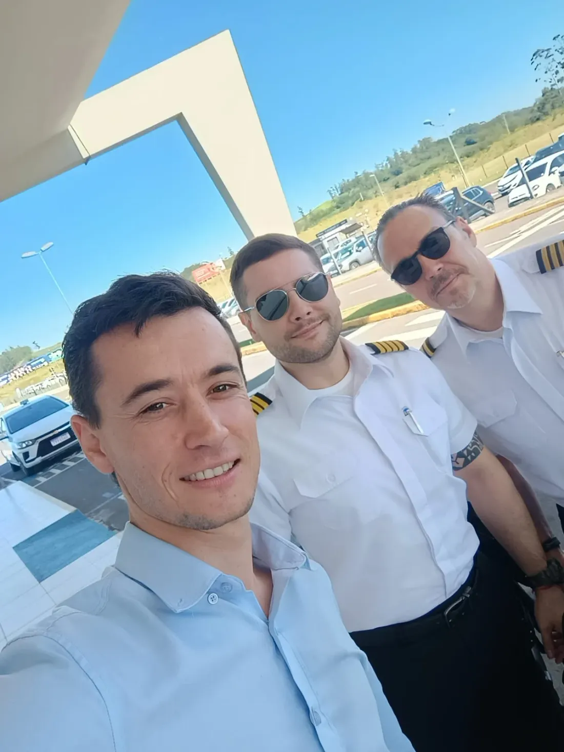 Selfie do motorista com dois pilotos de uniforme, na área de embarque de um aeroporto